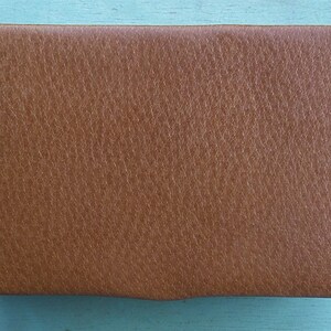 May include: A rectangular, brown leather case with a textured surface. The case has rounded corners and a slightly curved bottom edge. The leather appears to be a medium shade of brown, with a subtle grain pattern.
