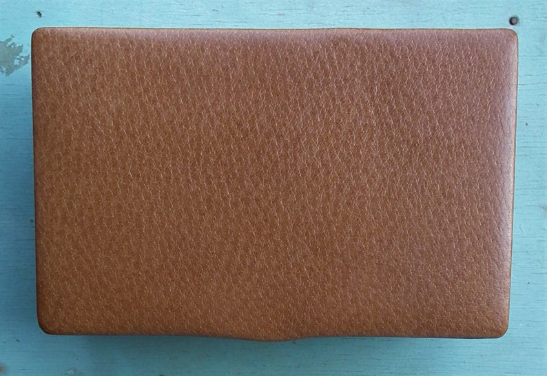 May include: A rectangular, brown leather case with a textured surface. The case has rounded corners and a slightly curved bottom edge. The leather appears to be a medium shade of brown, with a subtle grain pattern.