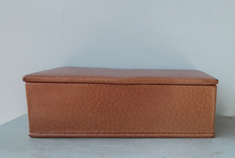 May include: A rectangular, brown leather box with a textured surface. The box has a hinged lid and is likely used for storage. The box is sitting on a light gray surface.
