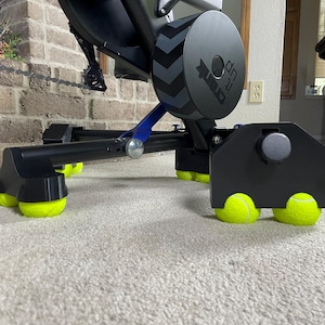 May include: A black exercise bike stand featuring a circular side housing with a chevron pattern and the text "up rank" printed on it. The base of the stand is supported by multiple bright neon yellow tennis balls attached to the feet.