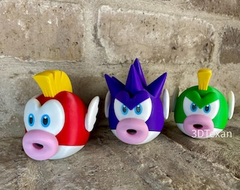 Puede incluir: Tres figuras de personajes de dibujos animados impresas en 3D de colores brillantes. Las figuras son rojas, moradas y verdes, cada una con vientre blanco, ojos azules y boca rosa. Cada una tiene una aleta o cresta amarilla. Las figuras se muestran sobre una superficie texturizada.