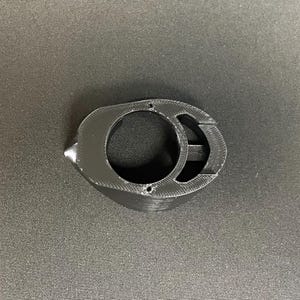 May include: A black 3D-printed component featuring a central circular opening, a secondary smaller oval aperture, and a pointed protrusion on one side. The object has a textured surface finish with visible layer lines from the manufacturing process.