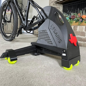 May include: A black Saris bike trainer with a red Texas-shaped emblem. The trainer has a black frame and yellow rollers. A black bicycle is mounted on the trainer, with the rear wheel in contact with the trainer's roller.