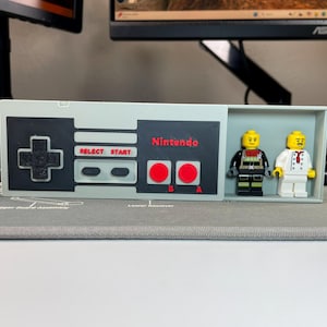 May include: A light gray box with a black and gray Nintendo controller design. The box contains two Lego figures. The controller design includes the word "Nintendo" in red, and red buttons. The box is on a gray surface.