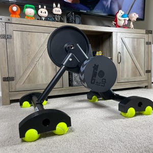 May include: Black exercise equipment with a large flywheel and the text "Crank It Up". The equipment has black legs with yellow tennis balls attached to the bottom. The equipment is on a carpeted floor.