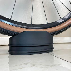 May include: A black bicycle wheel stand supporting a bicycle wheel. The wheel has a brown tire and black spokes. The stand is made of black plastic and has a grooved design.