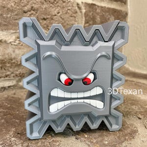 May include: A gray, 3D-printed Thwomp character from the Super Mario video game series. The character has an angry expression with red eyes and a wide, toothy grin. The text "3DTexan" is visible on the side.