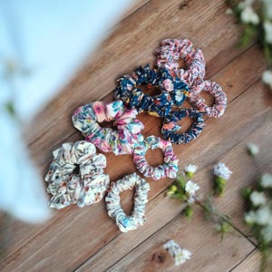 May include: A collection of floral patterned scrunchies in various colors, including pink, blue, and white, are arranged on a wooden surface. The scrunchies feature different floral designs and textures. Some scrunchies are clustered together, while others are spread out.