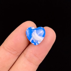 Pode incluir: Uma pedra em forma de coração com um padrão marmorizado azul e branco. A pedra é segurada entre dois dedos contra um fundo preto. O coração é liso e brilhante.