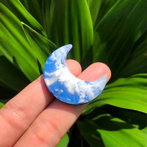 Könnte beinhalten: Ein halbmondförmiger Anhänger mit blauem Himmel und Wolkenmuster. Der Anhänger wird in einer Hand gehalten, mit einem grünen, blättrigen Hintergrund.