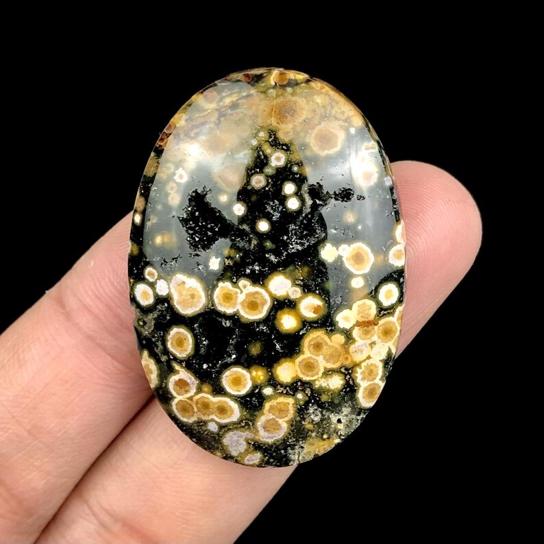 Pode incluir: Uma pedra preciosa oval com uma superf&iacute;cie polida. A pedra apresenta um padr&atilde;o de manchas e redemoinhos pretos, castanhos e creme. A gema &eacute; segurada em uma m&atilde;o contra um fundo preto. A pedra &eacute; provavelmente usada para joias ou fins decorativos.