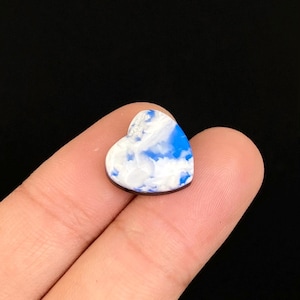 Pode incluir: Um pingente em forma de coração com um padrão azul e branco semelhante a nuvens. O pingente é segurado entre dois dedos contra um fundo preto. O pingente tem uma borda marrom escura.