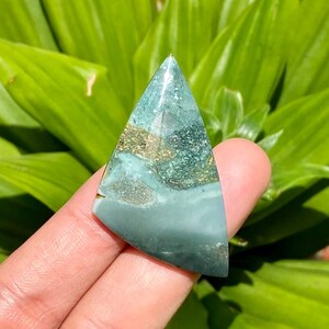 Op de afbeelding: Een driehoekige edelsteen met een mix van teal, goud en grijstinten. De gepolijste steen heeft een glad oppervlak en een puntige top. De achtergrond is een waas van groen gebladerte.