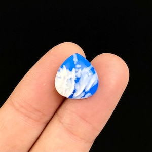 Pode incluir: Um pingente em forma de gota d'água azul e branco. O pingente tem uma base azul brilhante com padrões brancos em forma de nuvem. É segurado entre dois dedos em um fundo preto. O pingente tem aproximadamente 2 cm de comprimento.