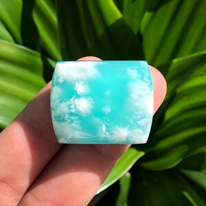 Pode incluir: Uma pedra preciosa retangular com bordas arredondadas, apresentando um fundo azul celeste com padrões brancos semelhantes a nuvens. A pedra é segurada em uma mão, com um fundo de folhagem verde.