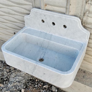 May include: A white marble sink with a backsplash and three holes for taps. The sink is rectangular and has a rounded edge. It is mounted on a wall.