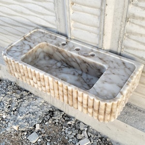 May include: A white marble bathroom sink with a unique fluted design around the edge. The sink has three holes for faucets.