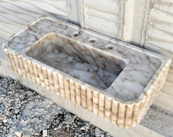 May include: A white marble bathroom sink with a unique fluted design around the edge. The sink has three holes for faucets.