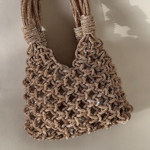 Puede incluir: Un bolso de mano tejido de color dorado, con un asa hecha de múltiples hebras. El cuerpo del bolso tiene un diseño único de tejido abierto. El asa está unida al bolso con secciones bien enrolladas. Un accesorio elegante.