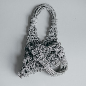 May include: A silver-toned handbag constructed from a complex, knotted rope-like material. The bag features a triangular silhouette with a single integrated handle and a textured, woven surface that creates a repeating geometric pattern.
