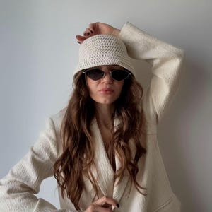 Puede incluir: Sombrero de pescador de ganchillo color crema y gafas de sol. El sombrero se combina con una blazer texturizada a juego. La imagen muestra un atuendo elegante en tonos neutros, perfecto para un look moderno.