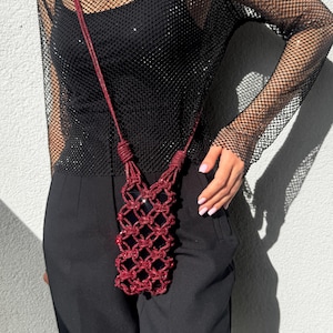 May include: A dark red, glittery macrame phone pouch featuring a knotted, open-weave grid design. The pouch includes a long, matching cord strap that is knotted at the top, allowing it to be worn as a crossbody accessory.