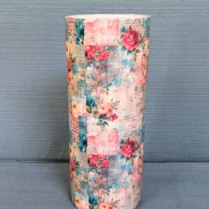 May include: A cylindrical tumbler featuring a collage-style print with pink roses, blue watercolor splashes, and faded script text. The design covers the entire exterior surface of the vessel, showcasing a blend of floral patterns and vintage writing.