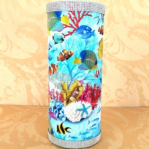 May include: Cylindrical decorative container featuring a vibrant underwater scene with colorful fish, coral, and sea life. The top and bottom rims are adorned with sparkling rhinestones. The container is a decorative item.