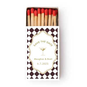 May include: A matchbox with a brown and white chequered pattern. The front of the box features a gold-outlined label with the words "Save the Date" and a cocktail glass illustration. The names Meaghan & Brett and the date 6.7.2025 are also printed on the label. The match heads are red.