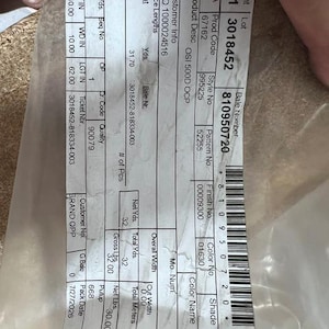 May include: A white rectangular product label printed with black text and a barcode, encased in clear plastic. The label displays technical data including Lot 3018452, Bale Number 810950720, Product Description OSI 5000D OCP, and various numerical codes.