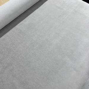 May include: A roll of light gray fabric with a smooth, uniform texture. The material is displayed in a continuous length, showing a consistent matte finish across the surface. The fabric is tightly wound at the top, creating a cylindrical shape.