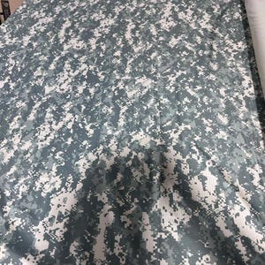 May include: A bolt of fabric featuring a digital camouflage pattern in shades of slate gray, white, and dark charcoal. The textile displays a pixelated, multi-tonal design across its surface, rolled onto a cylindrical core for storage and display.