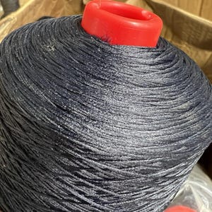 May include: A large spool of dark blue thread with a red plastic core. The thread is tightly wound, creating a textured surface. The image is a close-up, highlighting the thread's sheen and the core's bright color.