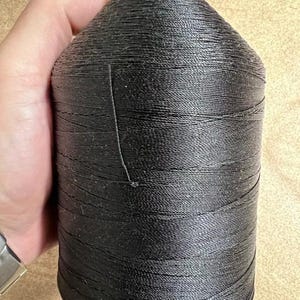 May include: A large, cylindrical spool of black thread featuring a smooth, tightly wound texture. The spool has a tapered top section with a flat, circular cap and a wider base, showcasing a consistent dark color throughout the entire length of the thread.