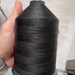 May include: A large, cylindrical spool of black thread featuring a tapered top and a flat base. The thread is tightly wound around the spool, creating a textured, horizontal pattern across the surface. The spool is constructed from a dark material.