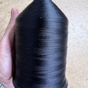 May include: A large, cylindrical spool of black thread featuring a smooth, tightly wound texture. The spool has a tapered top and a flat, circular base, designed for industrial or heavy-duty sewing applications, providing a consistent supply of thread.