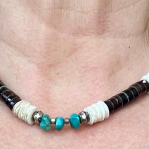 May include: A handcrafted necklace featuring alternating dark brown and white shell beads, accented with turquoise stones and silver-toned metal spacers. The necklace is displayed against a skin tone background.