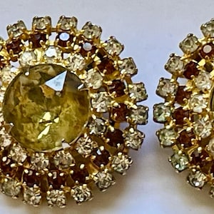 Puede incluir: Un par de pendientes de clip de tono dorado con piedras grandes, redondas, facetadas, de color verde amarillento rodeadas de pequeños diamantes de imitación transparentes y marrones.