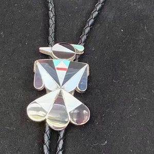 May include: A bolo tie featuring a central bird-shaped pendant inlaid with multicoloured stone and shell segments. The pendant is attached to a black braided leather cord that terminates in two long, tapered silver-toned metal tips with rounded ends.