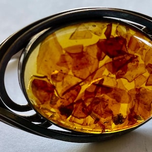 May include: A silver-toned bracelet with an oval amber centerpiece. The amber is filled with golden-brown inclusions, set within a swirling, openwork metal frame. The bracelet has a clasp closure.