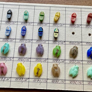 Puede incluir: Una colección de pequeños objetos decorativos, posiblemente cuentas o abalorios, en varios colores, incluyendo negro, azul, verde, amarillo y rosa. Cada pieza tiene un diseño único y está dispuesta en una cuadrícula. Los objetos están sobre una superficie beige.