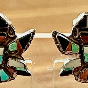 May include: A pair of silver-toned earrings with a Native American design. The earrings feature inlaid stones in black, white, turquoise, and red. The design is symmetrical and detailed, with a stylized bird or figure shape.