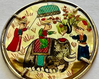 May include: Round pendant with a silver-colored frame. The pendant features a hand-painted scene of figures and an elephant. The figures are wearing colorful clothing. The background is a light yellow color with a pearlescent sheen.