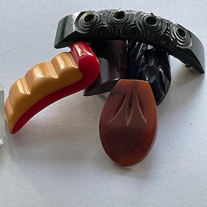 May include: A selection of vintage art deco jewellery pieces. Includes a dark green carved bracelet, a red and tan brooch, and a brown teardrop-shaped pendant with etched details. The pieces are arranged on a white surface.