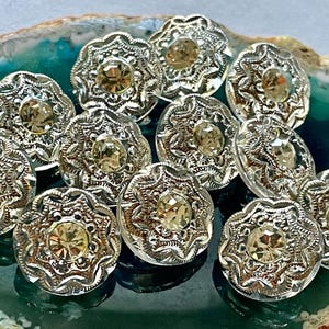 Puede incluir: Un conjunto de botones plateados con un diseño floral y una gema central de color amarillo claro. Los botones están dispuestos sobre una superficie verde y marrón. Estos botones decorativos son ideales para proyectos de costura y manualidades.