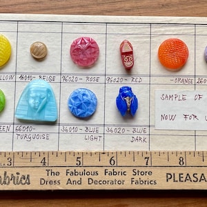 May include: A collection of vintage MoonGlow glass buttons in various colors and designs, including white, yellow, rose, red, orange, violet, and grey. The buttons are arranged on a grid with labels and a ruler.