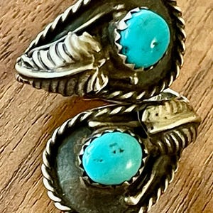 May include: A pair of silver-toned rings with a turquoise stone. The rings have a detailed design with a twisted rope border and ornate metalwork. The turquoise stones are round and set in a bezel setting.
