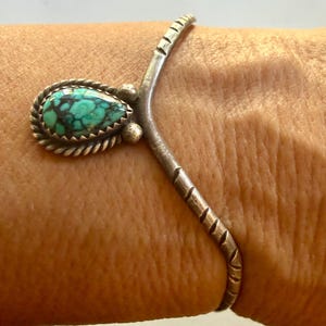May include: A silver cuff bracelet featuring a V-shaped design with etched diagonal lines along the band. A teardrop-shaped turquoise stone with dark matrix veining is set in a twisted silver rope bezel at the center of the V-shaped point.