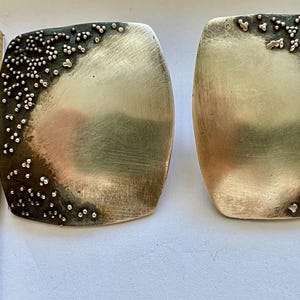 May include: A pair of silver square earrings with a textured, hammered finish. The earrings have a raised, textured design around the edges.