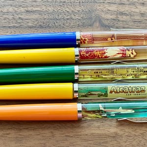 Könnte beinhalten: Fünf farbige Stifte mit transparenten Gehäusen, die Miniatur-Szenen zum Thema Reisen zeigen. Die Stifte haben blaue, gelbe, grüne und orangefarbene Körper. Ein Stift zeigt den Eiffelturm, ein anderer eine tropische Szene und ein weiterer Alcatraz.